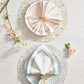 Two place settings on a white wood table, each atop a hand-crafted Stardust Placemat by Kim Seybert—one with a fan-fold napkin in a pink ring, the other triangular with silver—decorated with flowers.