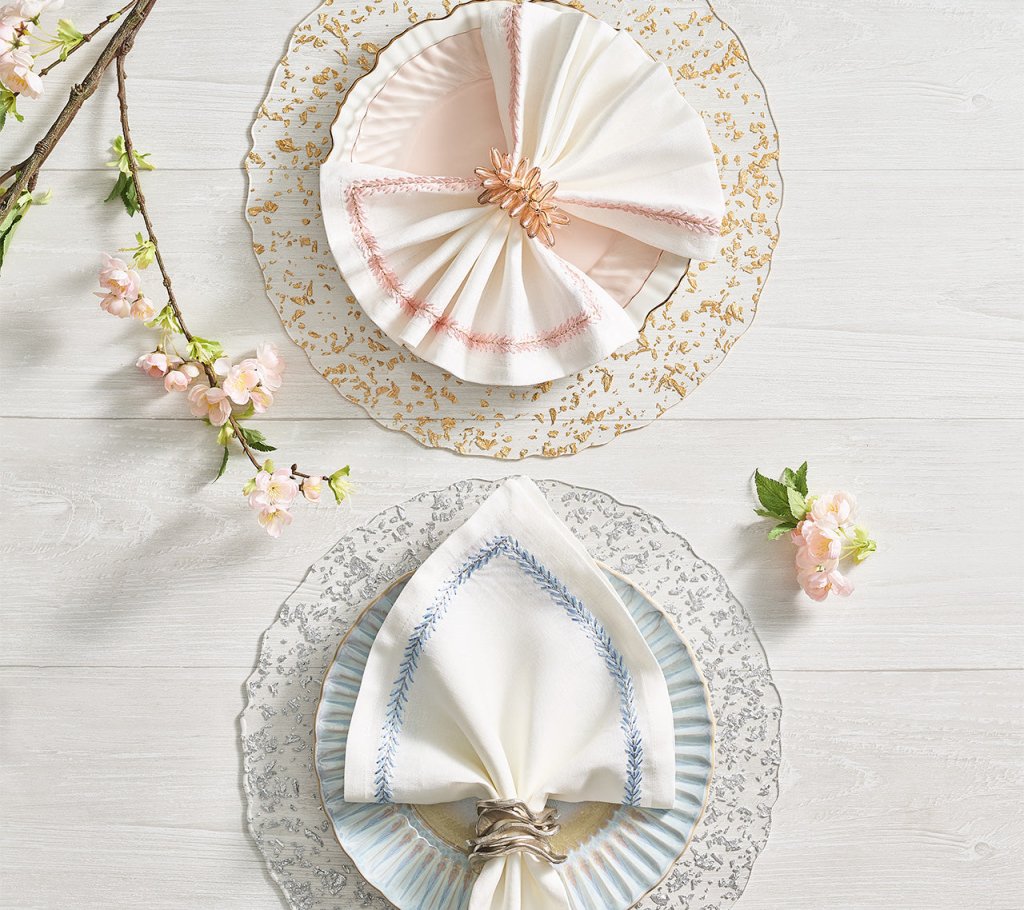Two place settings on a white wood table, each atop a hand-crafted Stardust Placemat by Kim Seybert—one with a fan-fold napkin in a pink ring, the other triangular with silver—decorated with flowers.