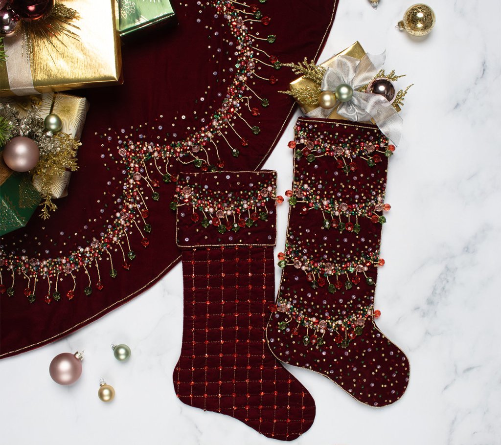 Two Kim Seybert Celestial Stockings in burgundy velvet with bead and sequin embellishments are shown on a marble surface alongside ornaments, wrapped gifts, and a coordinating tree skirt—an elegant holiday display.