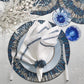 A table setting with a white napkin embroidered with blue leaves, blue glassware, silverware, a Kim Seybert Illusion Placemat, and a decorative white branch centerpiece.