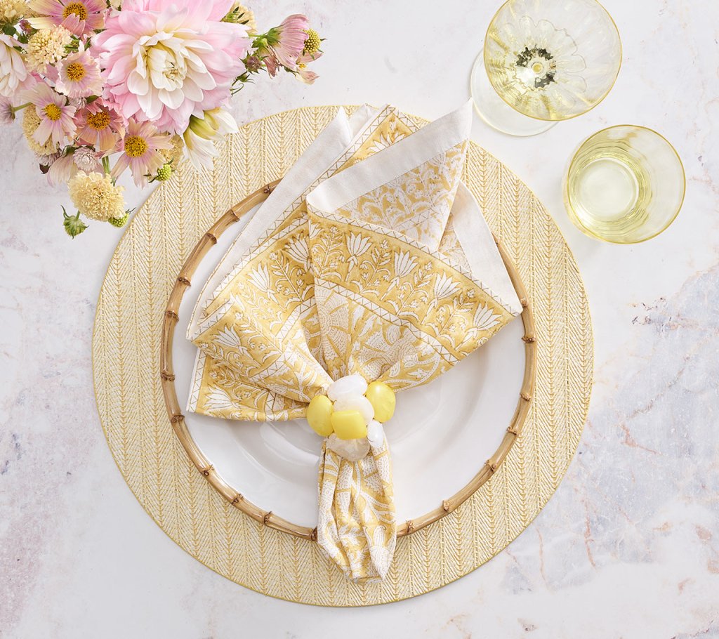 A table setting features a white plate on a yellow placemat, Kim Seybert's Provence Napkin Set with a fun pattern and napkin ring, two yellow glasses, and pink and white flowers on a matching tablecloth.