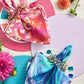 Two place settings with patterned napkins and Kim Seybert Fun Burst Napkin Rings featuring metallic beads, on textured placemats with pink and orange flowers.