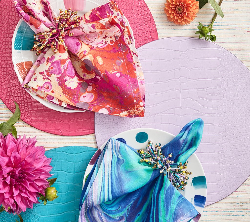 Two place settings with patterned napkins and Kim Seybert Fun Burst Napkin Rings featuring metallic beads, on textured placemats with pink and orange flowers.