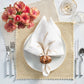 A table setting with a white plate, Kim Seybert Silver & Gold Stamped Reversible Placemat, folded white napkin in a holder, silver cutlery, two glasses, and a vase of pink flowers.