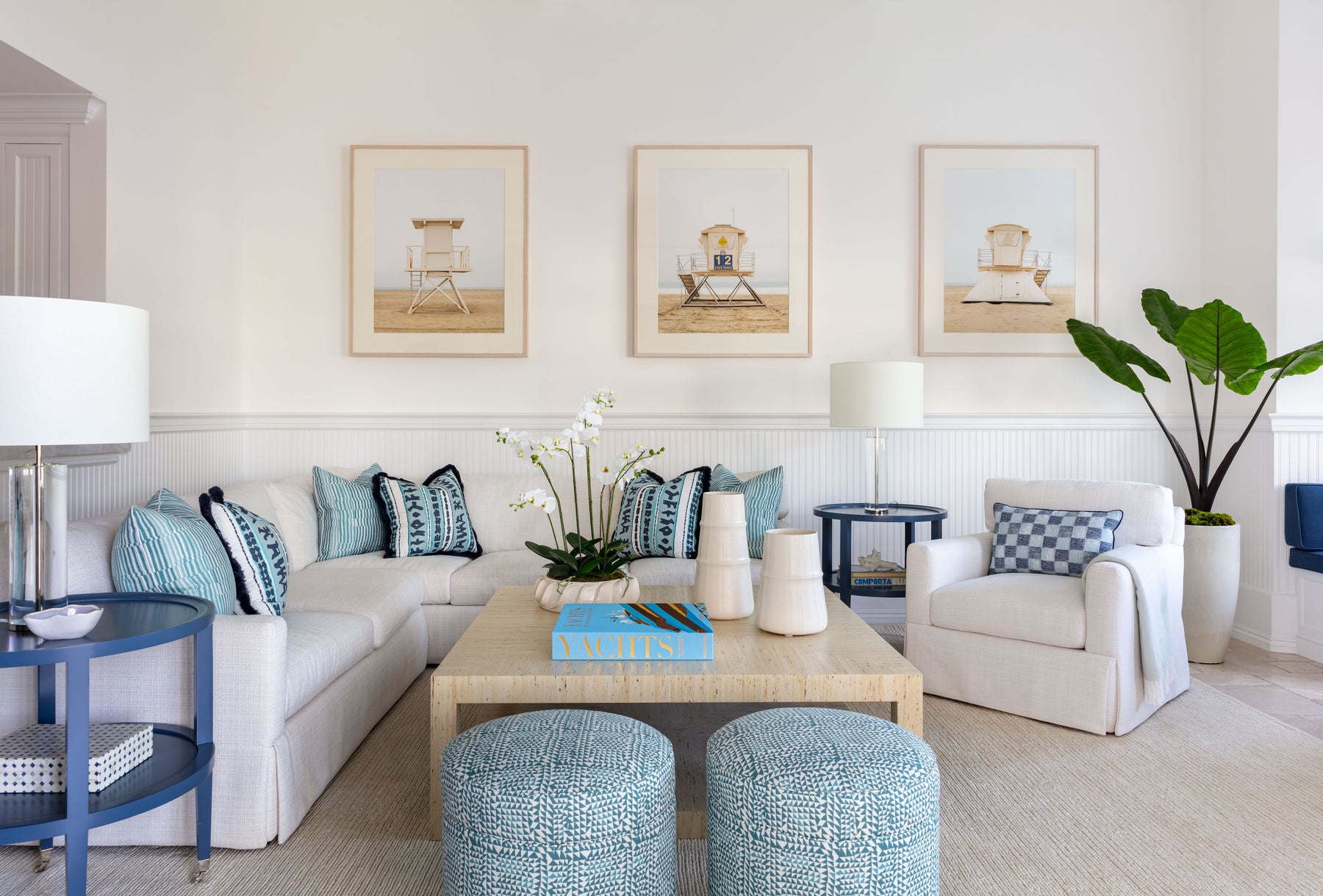 living area with large white sectional, blue patterned pillows, large neutral coffee table, coordinating upholstered blue stools and paintings on wall of beach lifeguard stands