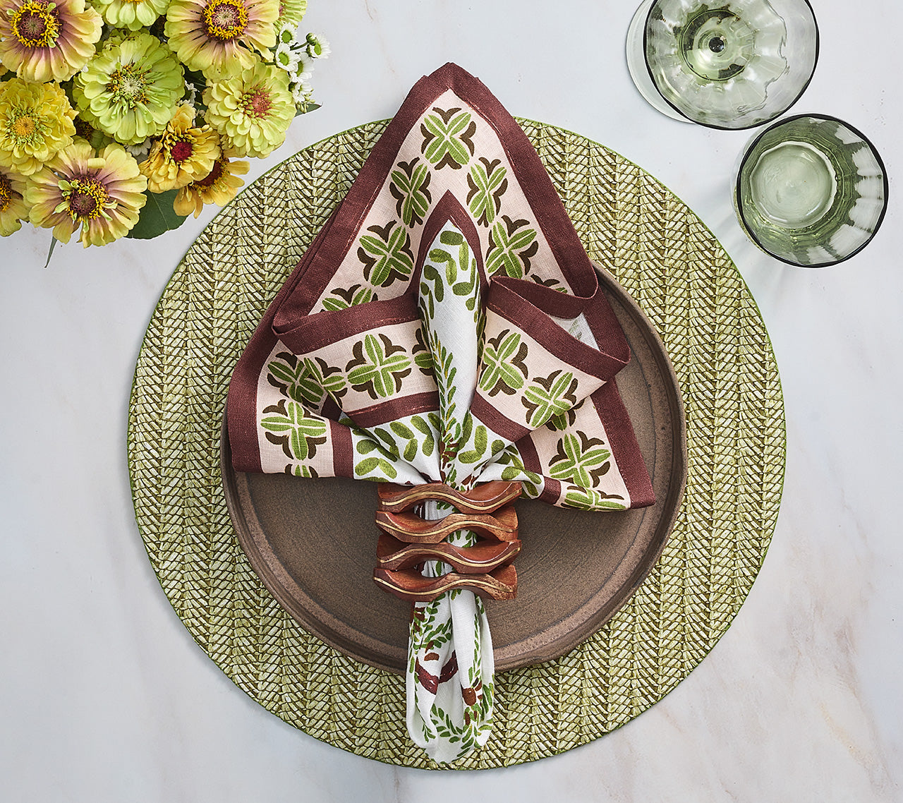 A dinner plate sits on the Kim Seybert Chevron Moss Placemat, accented by a folded napkin with ring, two green glasses, and yellow flowers in a vase atop a white table.