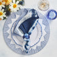 A blue and white tie-dye napkin with a blue ring rests on a patterned plate set atop the Kim Seybert Dream Weaver Placemat, beside a drink, a blue glass, and bright summer flowers—ideal for stylish entertaining.