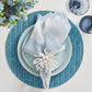 The Kim Seybert Watercolor Stripe Linen Collection elegantly sets a summer table with blue plates and a light blue napkin, accented by a white napkin ring on a textured blue placemat, accompanied by floral arrangements and two glass cups.