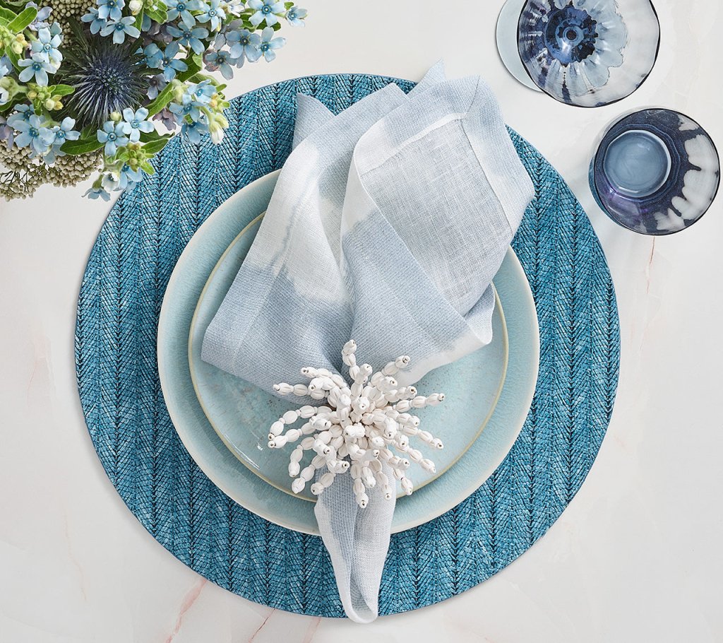 The Kim Seybert Watercolor Stripe Linen Collection elegantly sets a summer table with blue plates and a light blue napkin, accented by a white napkin ring on a textured blue placemat, accompanied by floral arrangements and two glass cups.