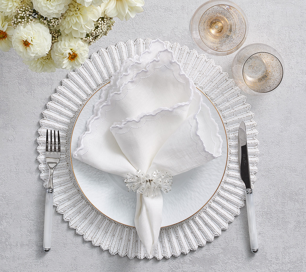 Elegant table setting featuring the Kim Seybert Arches Napkin Set, arranged with a white plate, placemat, napkin ring, silverware, two glasses, and white flowers atop a light gray surface.