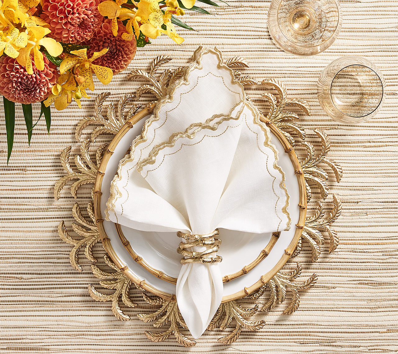 A white plate with a gold rim is topped with a white napkin in the Kim Seybert Gold Bamboo Napkin Ring, set on a decorative gold placemat with yellow and orange flowers nearby.