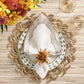 A formal table is set with a decorative gold placemat, white plate with green accents, Kim Seybert's Frame Napkin Set folded with gold trim and floral ring, metallic touches, two glasses, and a yellow-red floral centerpiece.