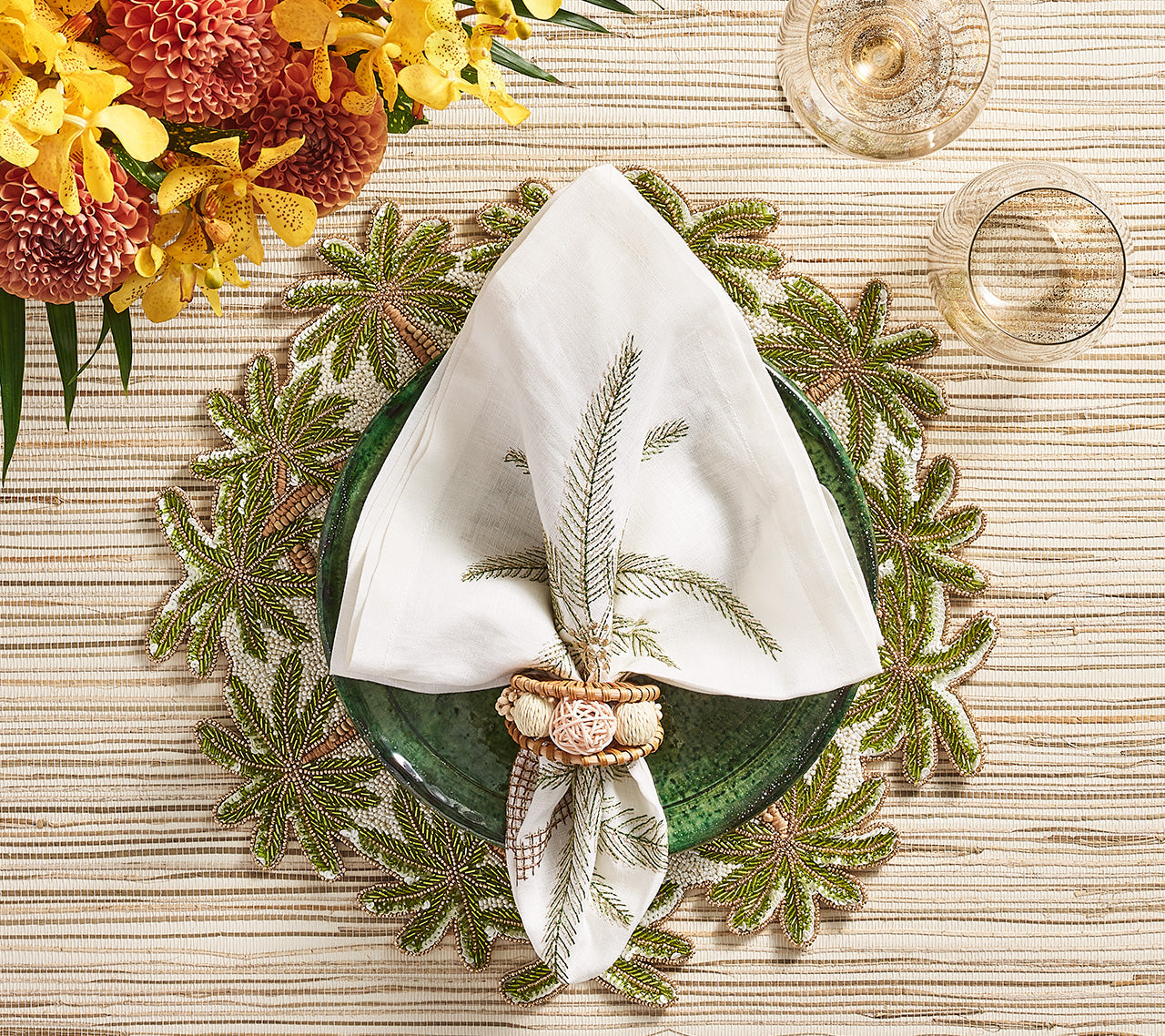 A green glass plate with a white napkin and beaded palm tree napkin ring rests on a Kim Seybert Oasis Placemat, perfect for tropical entertaining, alongside two glasses and a nearby floral arrangement.