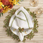 A white plate with a green embroidered napkin, held by the Kim Seybert Palm Coast Napkin Ring (Set of 4), sits on a leafy placemat beside wine glasses and flowers for nature-inspired table decor.