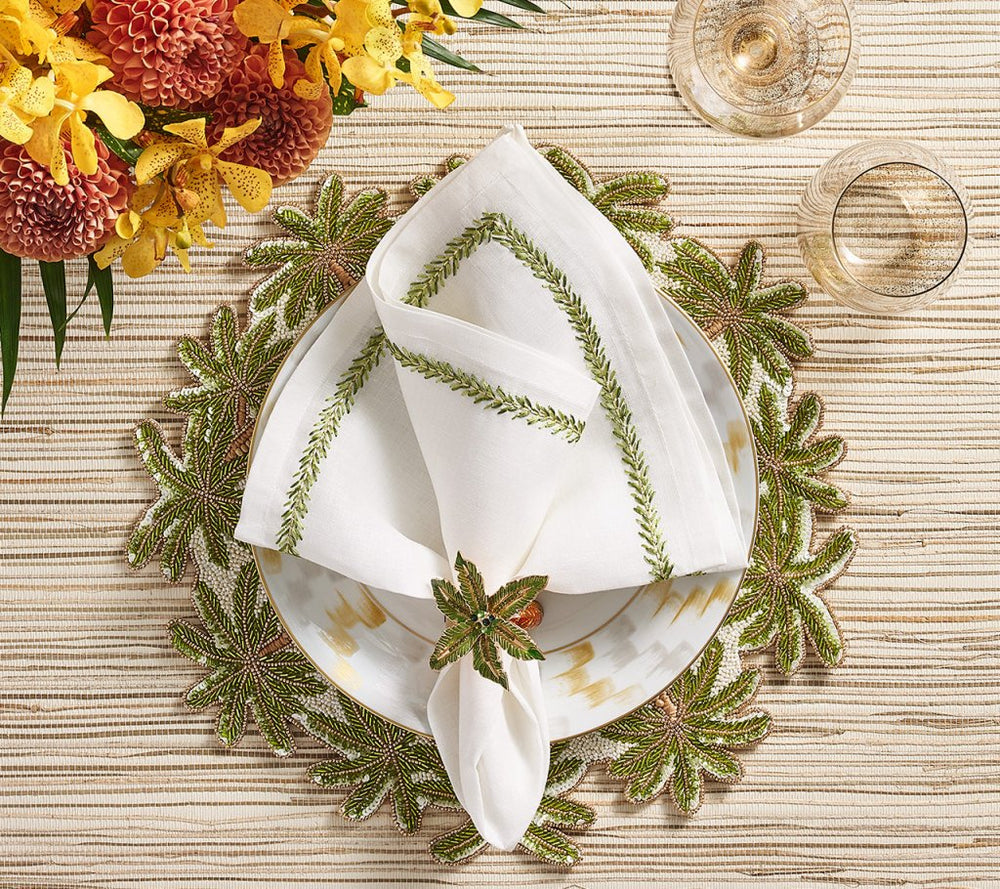 A white plate with a green embroidered napkin, held by the Kim Seybert Palm Coast Napkin Ring (Set of 4), sits on a leafy placemat beside wine glasses and flowers for nature-inspired table decor.