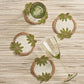 The Kim Seybert Oasis Coasters Set features four round, beaded coasters with green leaf designs—ideal for cocktail parties. Two green glasses, one upright and one tipped over, rest atop these stylish coasters on a striped surface.