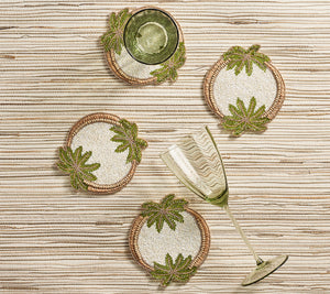 The Kim Seybert Oasis Coasters Set features four round, beaded coasters with green leaf designs—ideal for cocktail parties. Two green glasses, one upright and one tipped over, rest atop these stylish coasters on a striped surface.