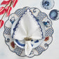 An elegant place setting features a blue and white patterned plate, the Kim Seybert Arches Napkin Set with embroidered edges, a napkin ring, two glasses, and pink flowers on a light surface.