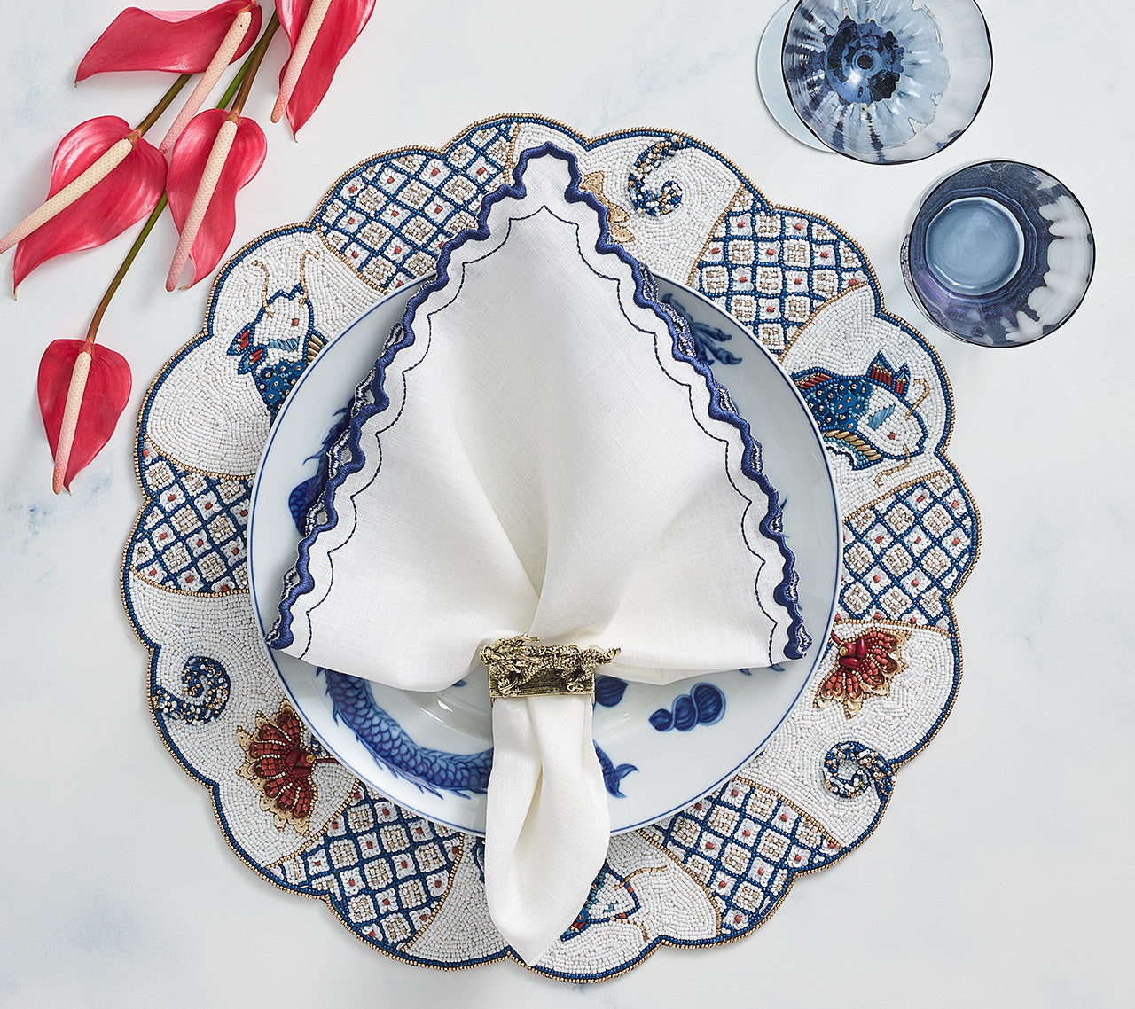 An elegant place setting features a blue and white patterned plate, the Kim Seybert Arches Napkin Set with embroidered edges, a napkin ring, two glasses, and pink flowers on a light surface.