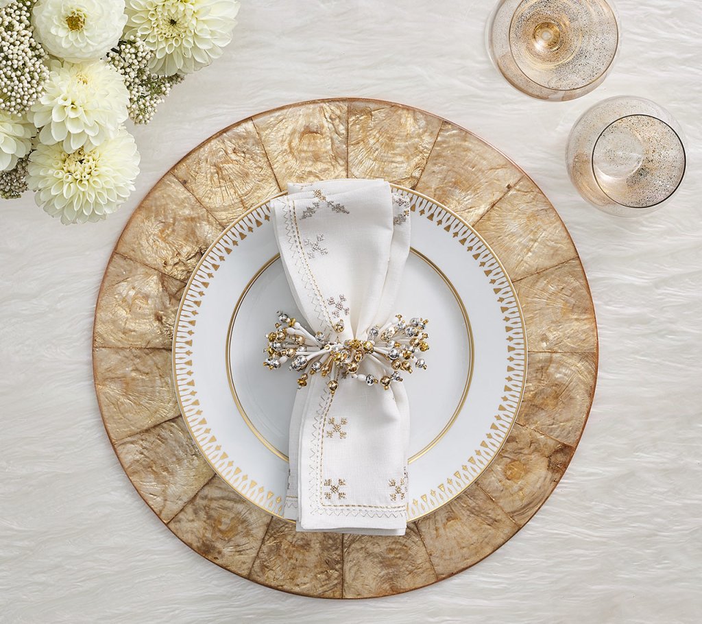 A formal place setting features a gold-rimmed plate, a white napkin with metallic beads in a Kim Seybert Fun Burst Napkin Ring, on a textured charger, accented by flowers and two glasses.