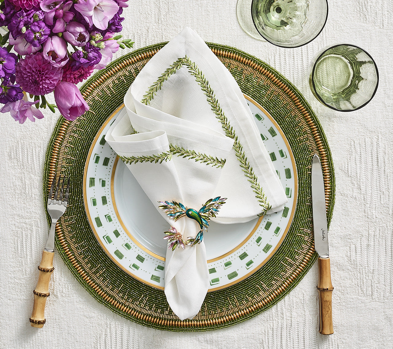 A white napkin with green trim, held by a butterfly ring, sits on patterned plates atop the Enamor Placemat by Kim Seybert. Bamboo cutlery, green glasses, and a purple floral arrangement create a charming spring setting.