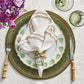 A beautiful table setting features a green woven placemat, a white plate with a delicate green leaf pattern, and an elegantly folded napkin secured by the Kim Seybert Gold Flux Napkin Ring. The look is completed with bamboo-handled fork and knife, two green glasses, and vibrant purple flowers for a touch of sophistication.