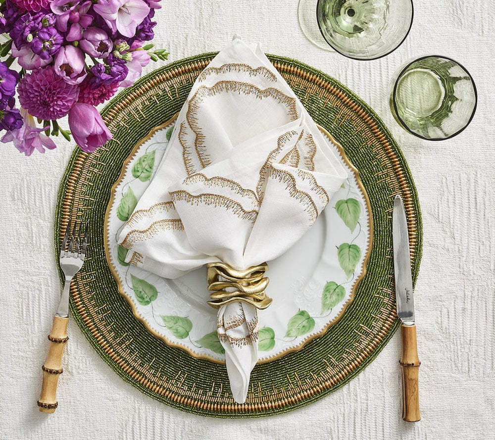 A beautiful table setting features a green woven placemat, a white plate with a delicate green leaf pattern, and an elegantly folded napkin secured by the Kim Seybert Gold Flux Napkin Ring. The look is completed with bamboo-handled fork and knife, two green glasses, and vibrant purple flowers for a touch of sophistication.