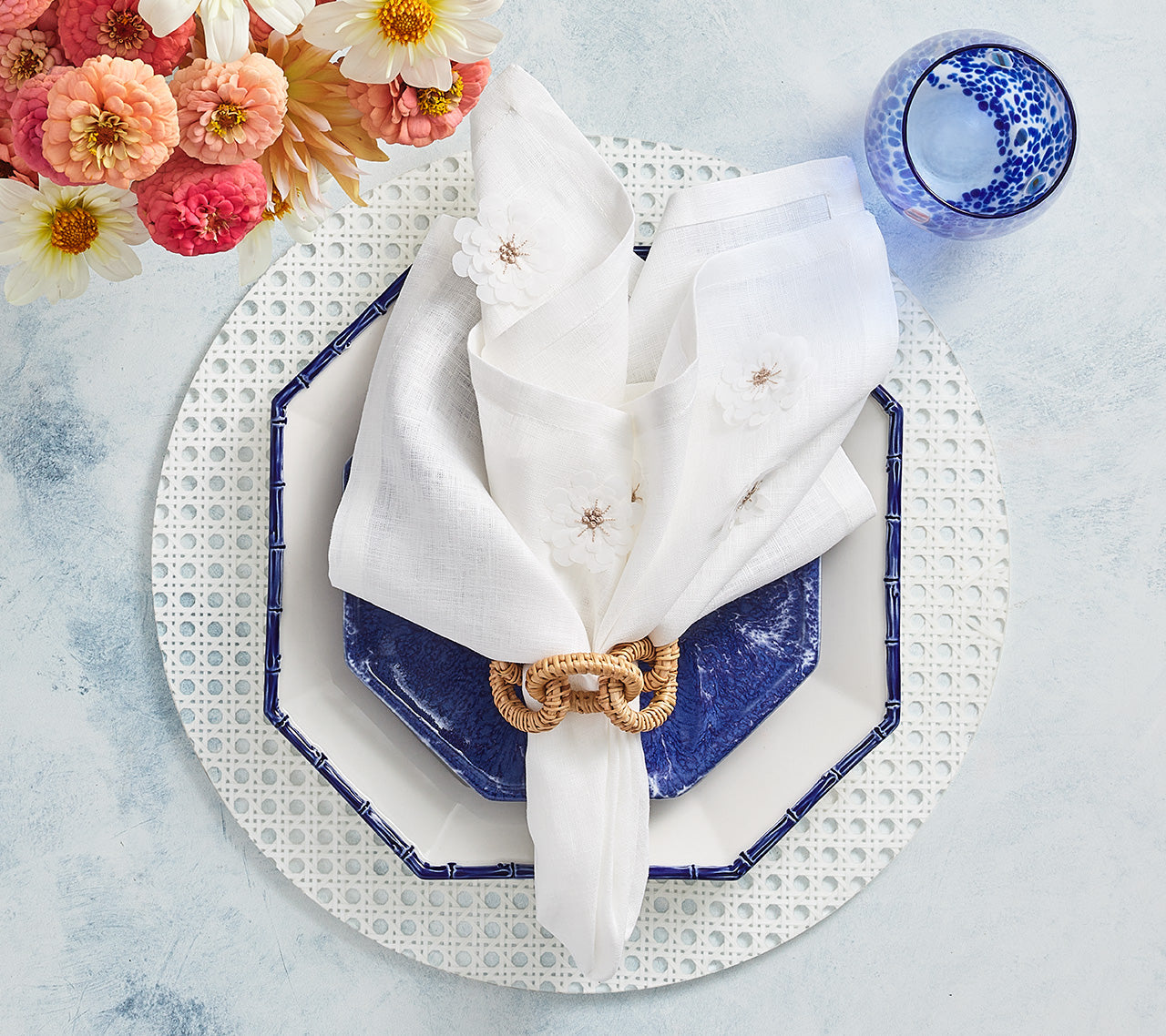 A blue glass and plate with a white napkin in ring rest on the Kim Seybert Reed Placemat beside pink and yellow flowers—an ideal setup for outdoor entertaining on a light blue surface.