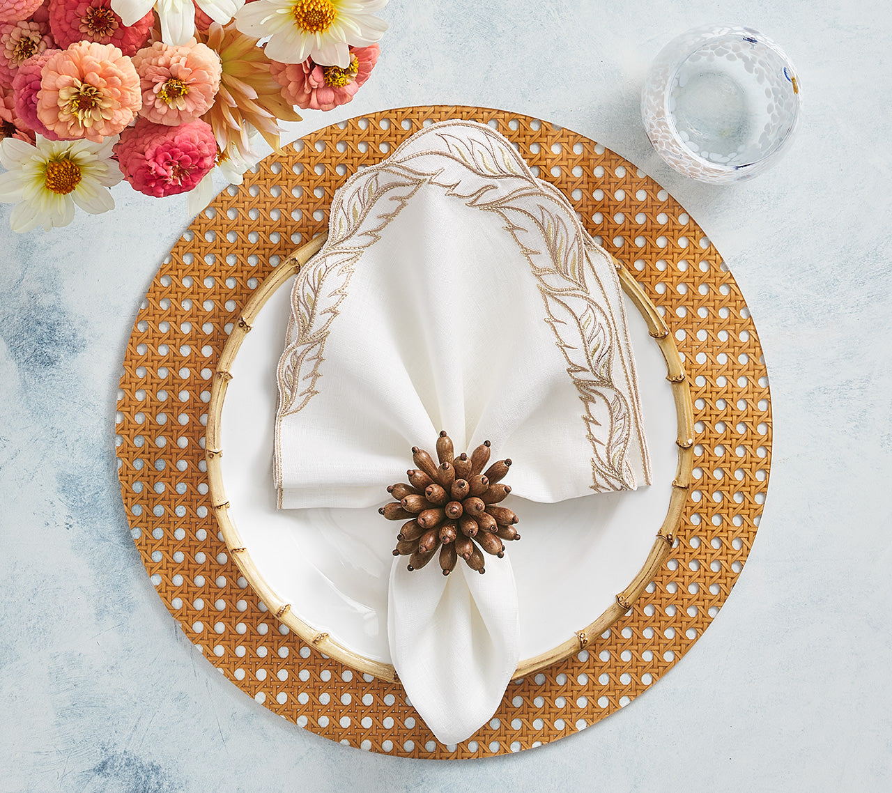 The Kim Seybert Reed Placemat holds a white plate with a gold rim, topped by a leaf-patterned napkin and ring—ideal for outdoor entertaining. A glass of water and pink flowers complete the setting.