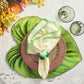 A table setting featuring the Kim Seybert Tropicana Placemat, a brown woven plate, a napkin with yellow stripes and green trim, green glassware, and assorted tropical fruits on the side.
