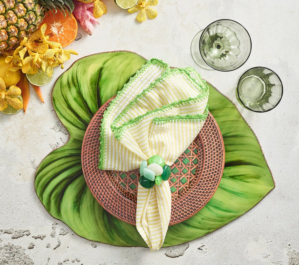 A table setting featuring the Kim Seybert Tropicana Placemat, a brown woven plate, a napkin with yellow stripes and green trim, green glassware, and assorted tropical fruits on the side.