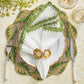 A formal table setting featuring Kim Seybert’s Shell Edge Napkin Set, a white plate, green and gold leaf placemat, knife and fork, two gold-rimmed glasses, and a linen napkin with raffia border in a gold napkin ring—ideal for chic bohemian tables.