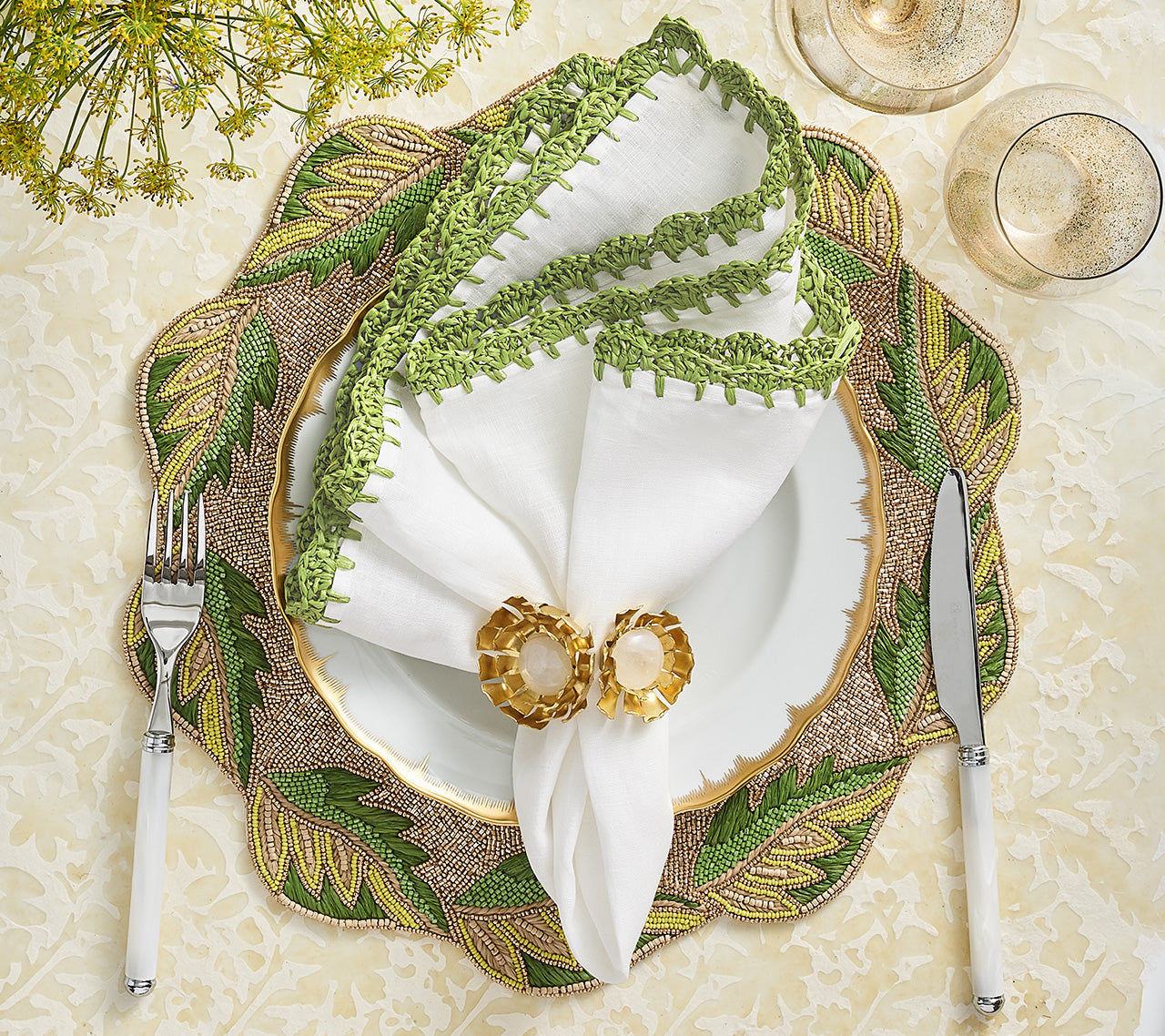 A formal table setting featuring Kim Seybert’s Shell Edge Napkin Set, a white plate, green and gold leaf placemat, knife and fork, two gold-rimmed glasses, and a linen napkin with raffia border in a gold napkin ring—ideal for chic bohemian tables.