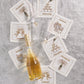 A bottle of champagne, two glasses, and the Kim Seybert Cheers Cocktail Napkin Set featuring six embroidered linen napkins are arranged on a silvery textured surface.