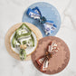 Three place settings with round placemats in blue, gold, and terracotta each feature a Kim Seybert Botanica Napkin Set and matching decorative napkin ring, all arranged on a marble surface.