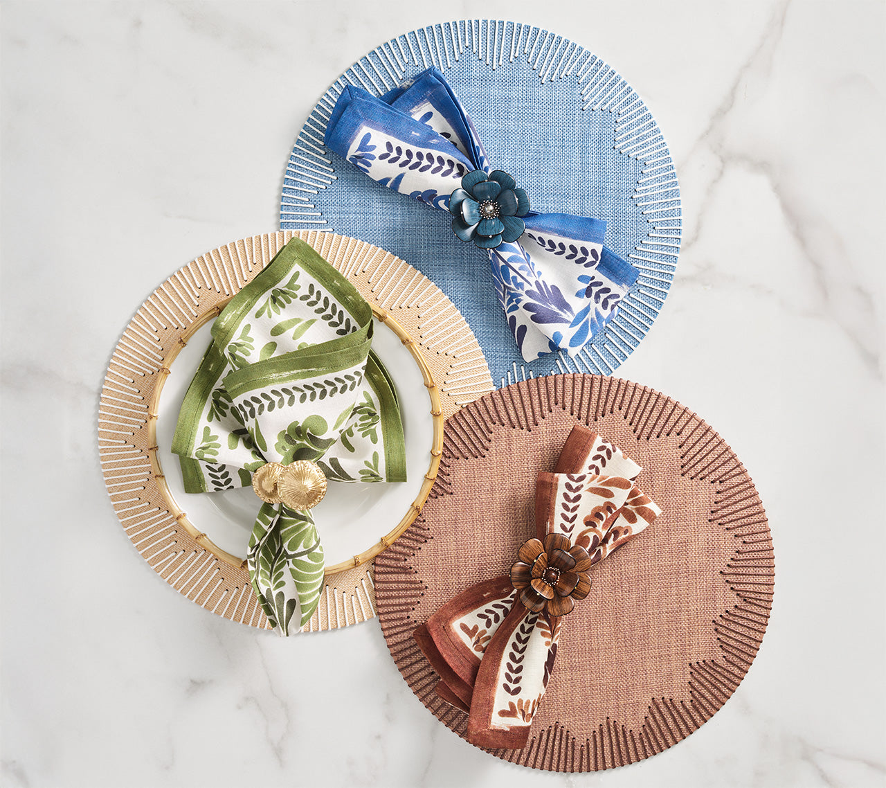 Three place settings with round placemats in blue, gold, and terracotta each feature a Kim Seybert Botanica Napkin Set and matching decorative napkin ring, all arranged on a marble surface.