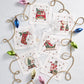 Six Christmas-themed coasters are arranged in a circle around a gold string of colorful bulb ornaments on a white surface. At the center are Kim Seybert Holly Jolly Cocktail Napkins, along with a wine glass and a cocktail glass.