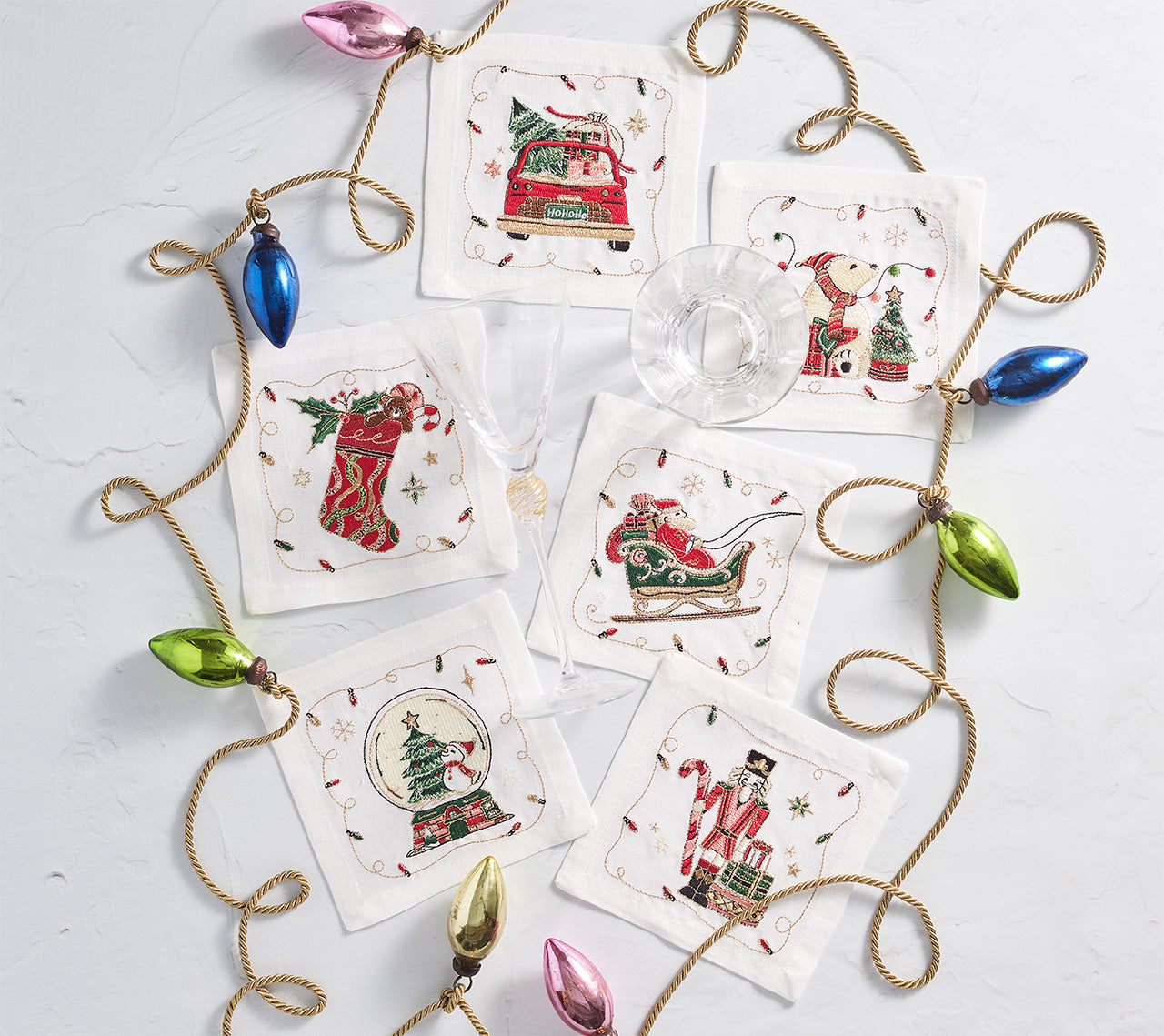 Six Christmas-themed coasters are arranged in a circle around a gold string of colorful bulb ornaments on a white surface. At the center are Kim Seybert Holly Jolly Cocktail Napkins, along with a wine glass and a cocktail glass.