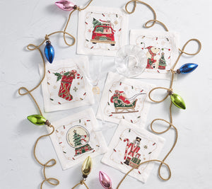 Six Christmas-themed coasters are arranged in a circle around a gold string of colorful bulb ornaments on a white surface. At the center are Kim Seybert Holly Jolly Cocktail Napkins, along with a wine glass and a cocktail glass.