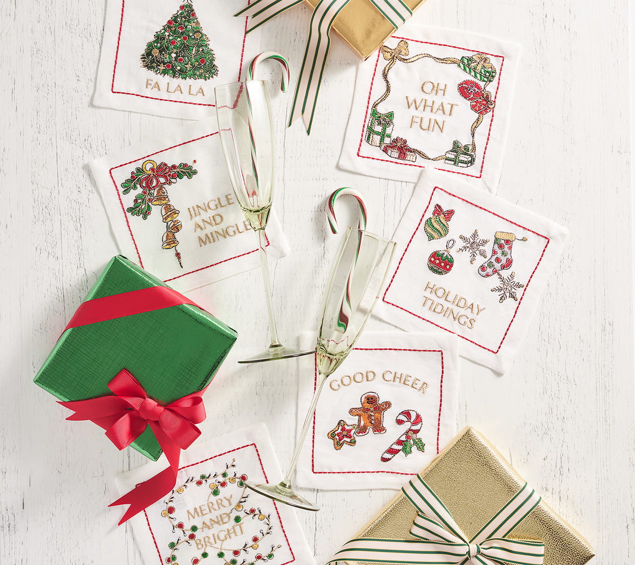 Two wrapped gifts, two champagne flutes with candy canes, and a Kim Seybert Merry and Bright Cocktail Napkin Set arranged on a white wooden surface—an ideal choice for a cheerful hostess gift.