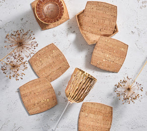 A set of square, heat-resistant coasters from the Kim Seybert Whistler Coaster Set with Caddy, a cork bowl, a gold-rimmed glass, and dried allium stems displayed on a textured white surface.