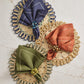 Three round woven placemats are paired with blue, orange, and green cloth napkins, each secured by Kim Seybert’s Bonton Napkin Rings featuring marbleized sapphire beads, all arranged on a light marble surface.
