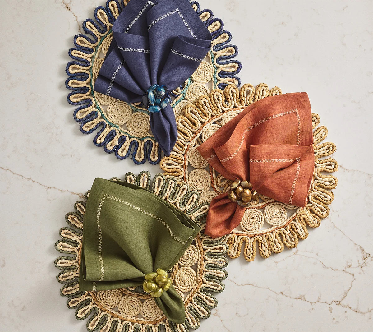 Three round woven placemats are paired with blue, orange, and green cloth napkins, each secured by Kim Seybert’s Bonton Napkin Rings featuring marbleized sapphire beads, all arranged on a light marble surface.