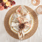 A table setting featuring Kim Seybert’s Giverny Napkin Set with a brown napkin ring, a gold beaded charger, white plate, and crystal glassware on light wood, accented by flowers.