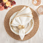 Table setting featuring a gold and white plate, white napkin with gold trim and shell-shaped ring, gold-toned glasses, flowers, all atop the Kim Seybert Emberfall Placemat on a light wood table.