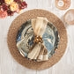 Table setting with a marbled plate on a textured placemat, beige napkin accented by the Kim Seybert Bonton Napkin Ring, two glasses, and a bouquet of flowers on a light wood surface.