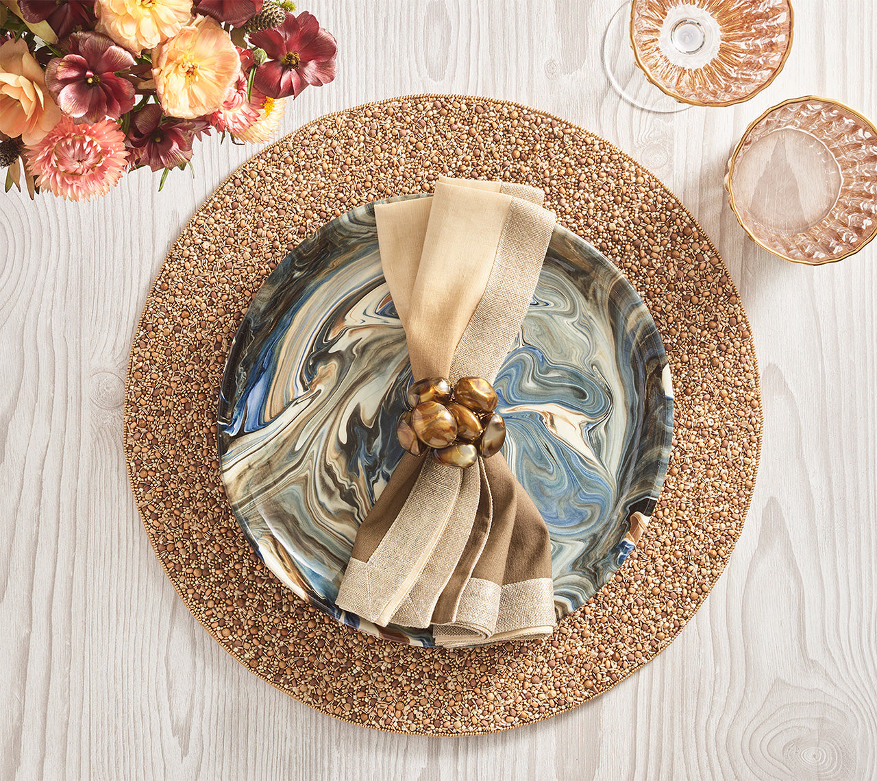 Table setting with a marbled plate on a textured placemat, beige napkin accented by the Kim Seybert Bonton Napkin Ring, two glasses, and a bouquet of flowers on a light wood surface.