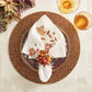 A fall-themed place setting features a beaded charger, patterned plate, embroidered napkin with a Kim Seybert Cornucopia Napkin Ring, and two amber glasses on a light surface accented with foliage.