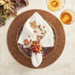 White napkin with embroidered autumn leaves held by a Kim Seybert Cornucopia Napkin Ring featuring faux pumpkins, berries, and foliage, resting on a dark plate and beaded charger for fall gatherings.