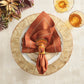 A rust-colored cloth napkin held by a metallic Kim Seybert Turkey Trot Napkin Ring, resting on a white dinner plate and a textured charger for autumn decor.